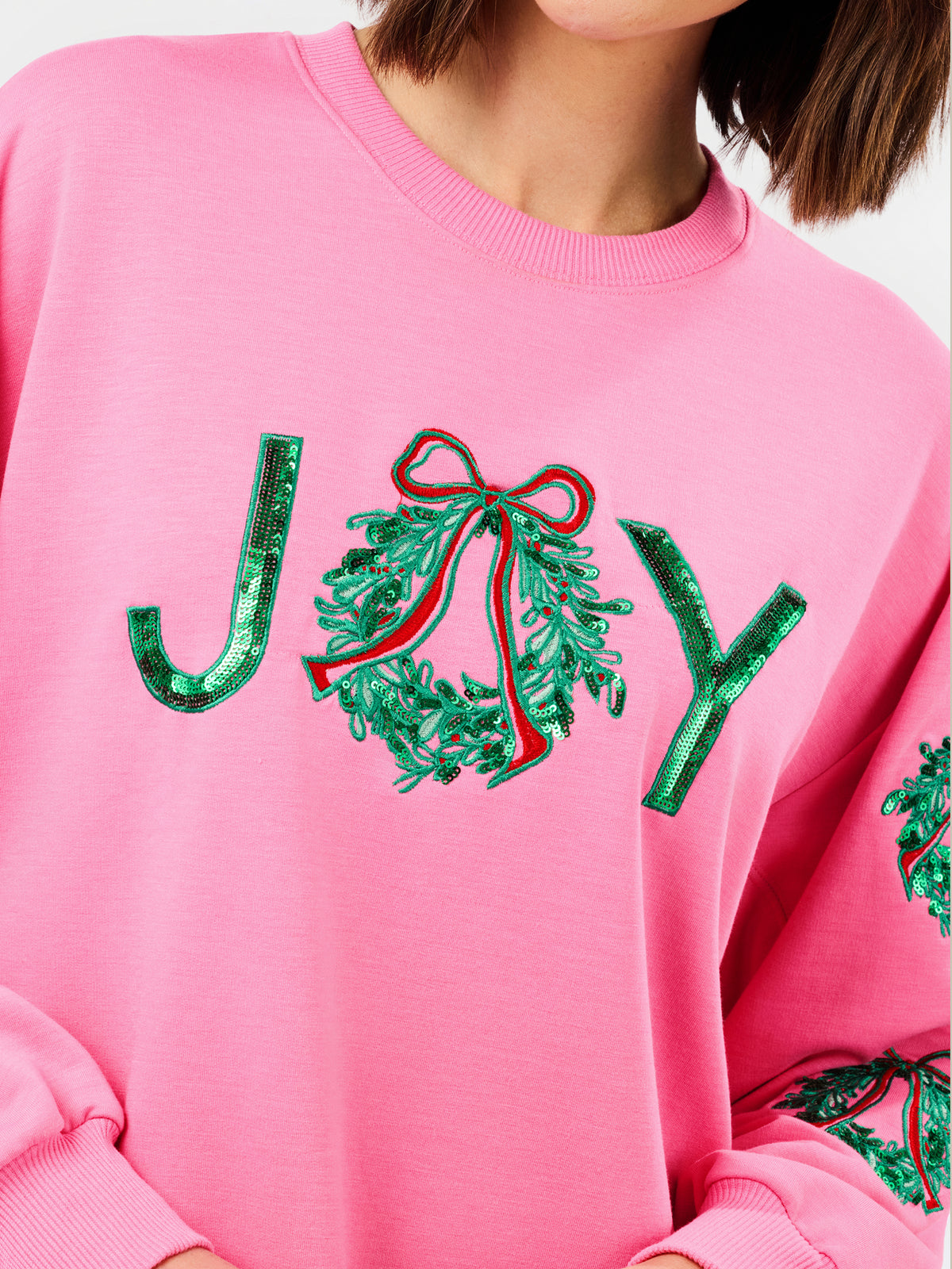 A close-up of a pink crewneck sweatshirt featuring a sequined 'JOY' graphic on the chest, with the 'O' represented by a green sequin wreath with a red bow.