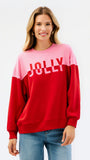 A woman wearing a color-blocked crewneck sweatshirt with a pink upper section and red lower section, featuring the word 'JOLLY' printed in white across the chest, paired with blue jeans against a plain white background.