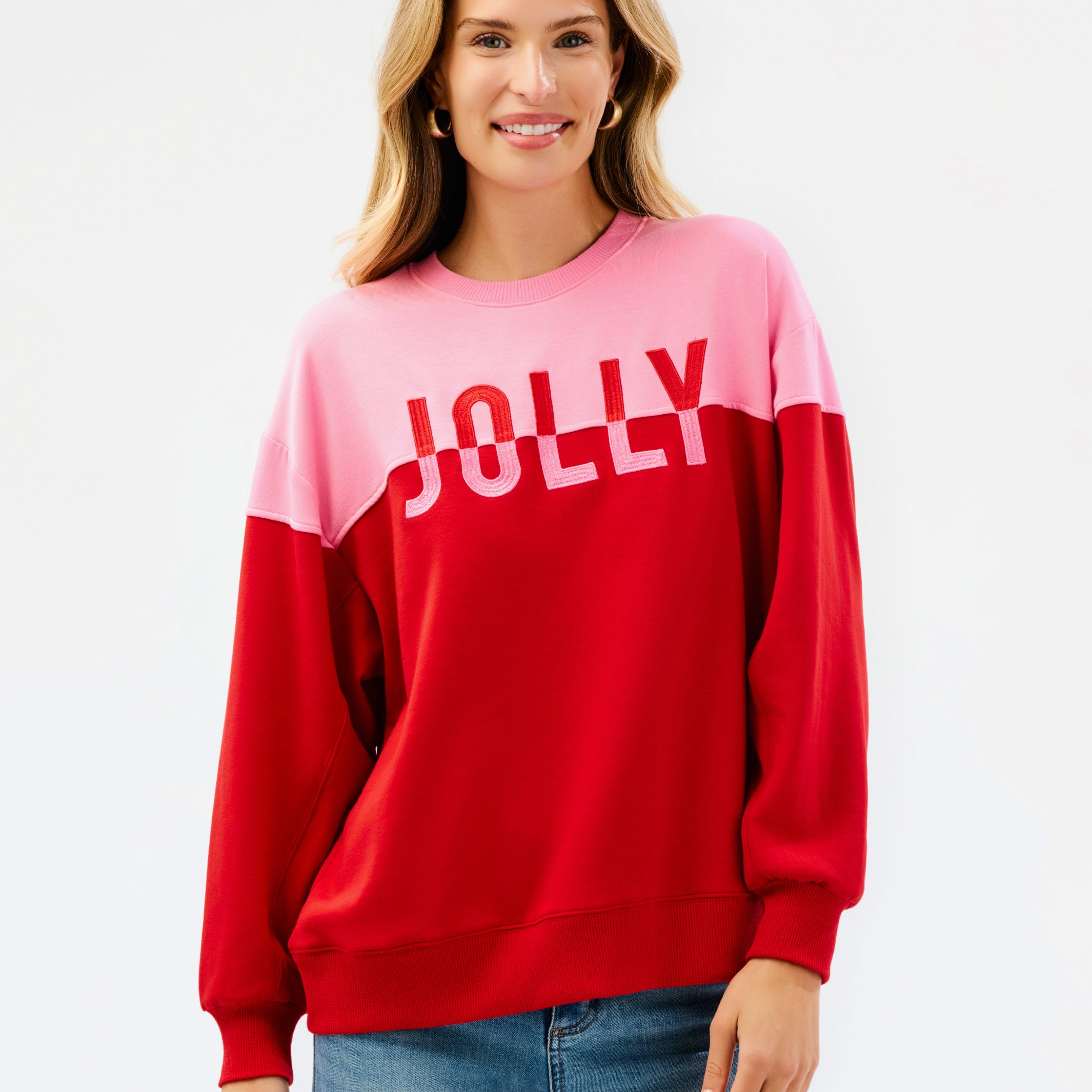 A woman wearing a color-blocked crewneck sweatshirt with a pink upper section and red lower section, featuring the word 'JOLLY' printed in white across the chest, paired with blue jeans against a plain white background.