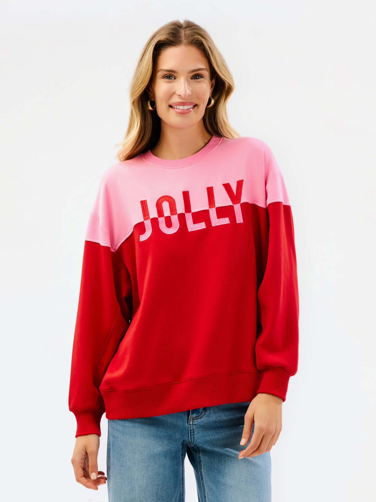 A woman wearing a color-blocked crewneck sweatshirt with a pink upper section and red lower section, featuring the word 'JOLLY' printed in white across the chest, paired with blue jeans against a plain white background.