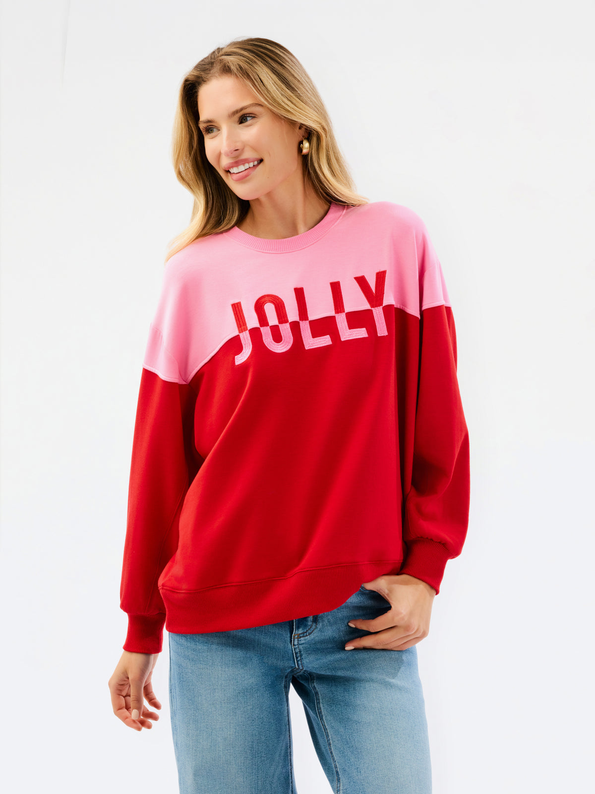 A woman wearing a color-blocked crewneck sweatshirt with a pink upper section and red lower section, featuring the word 'JOLLY' printed in white across the chest, paired with blue jeans against a plain white background.