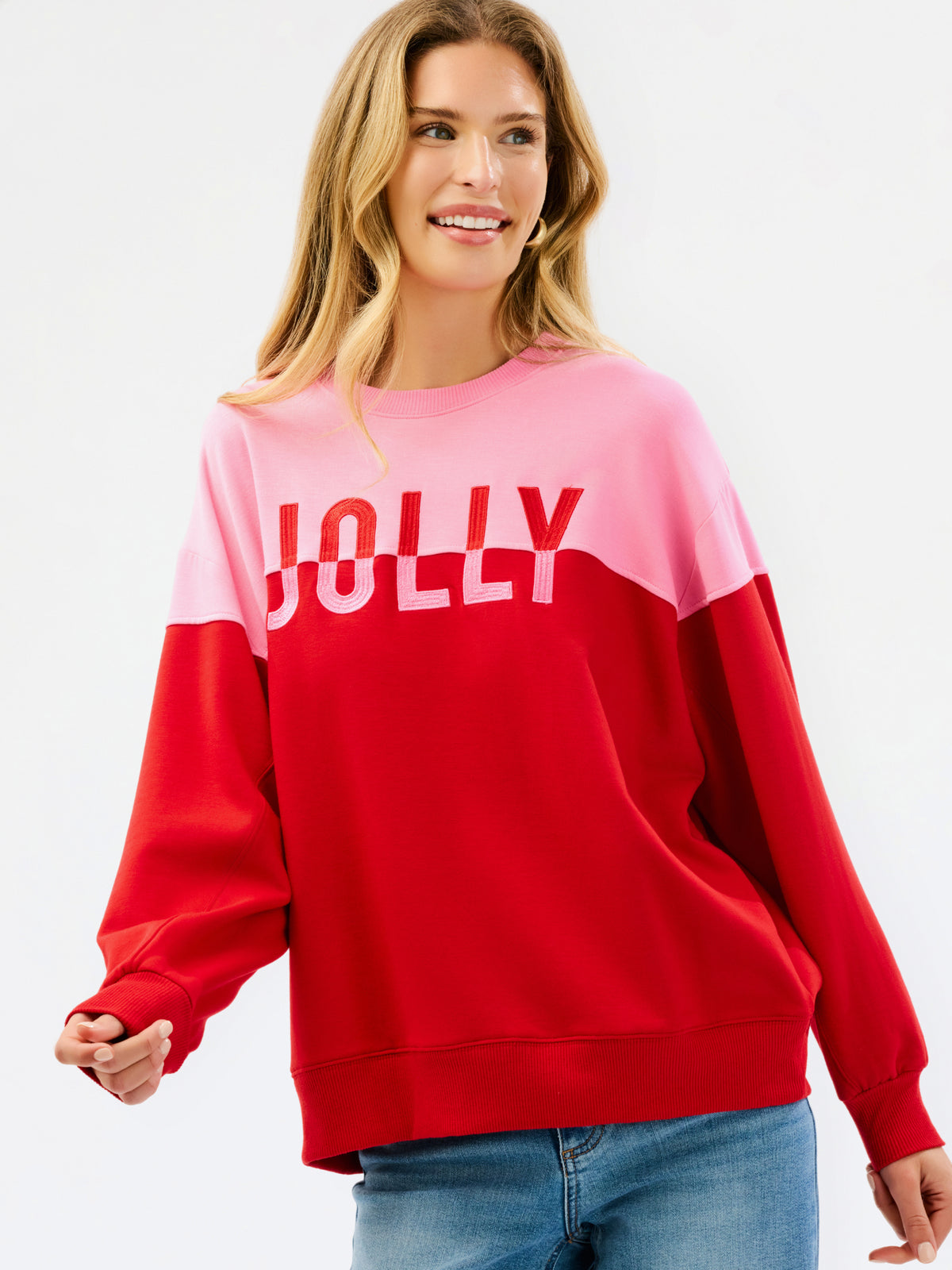 A woman wearing a color-blocked crewneck sweatshirt with a pink upper section and red lower section, featuring the word 'JOLLY' printed in white across the chest, paired with blue jeans against a plain white background.