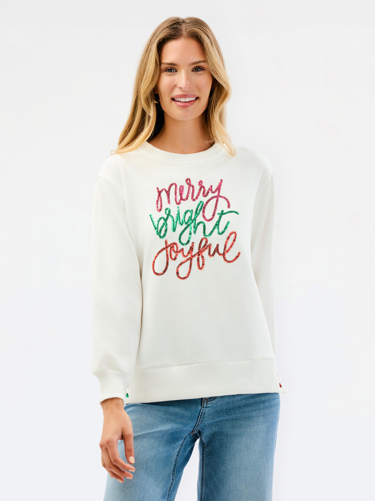 A woman wearing a cream-colored crewneck sweatshirt with 'Merry Bright Joyful' written in red and green cursive text on the front, paired with blue jeans, against a plain white background.