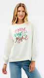 A woman wearing a cream-colored crewneck sweatshirt with 'Merry Bright Joyful' written in red and green glitter-style lettering on the front, paired with blue jeans, against a plain white studio background.
