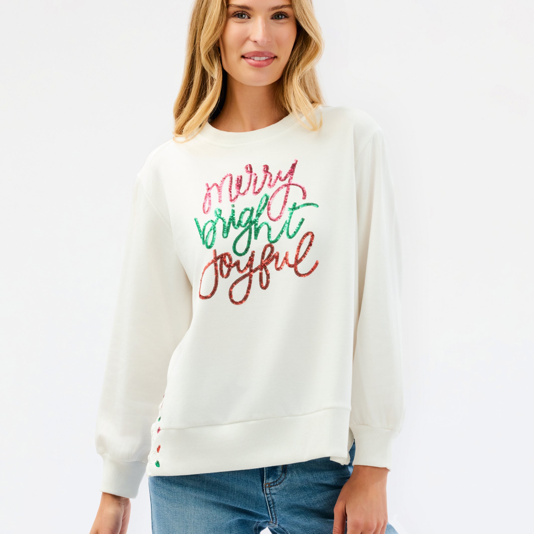 A woman wearing a cream-colored crewneck sweatshirt with 'Merry Bright Joyful' written in red and green glitter-style lettering on the front, paired with blue jeans, against a plain white studio background.