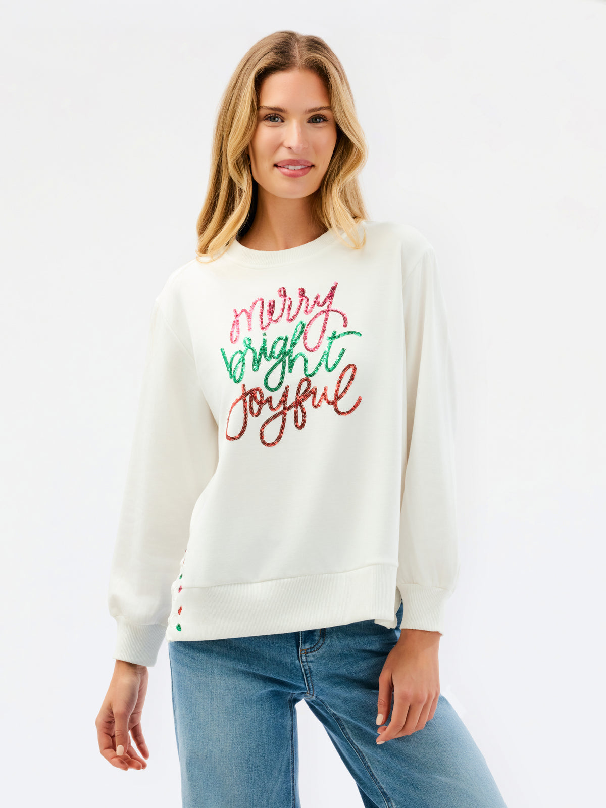 A woman wearing a cream-colored crewneck sweatshirt with 'Merry Bright Joyful' written in red and green glitter-style lettering on the front, paired with blue jeans, against a plain white studio background.