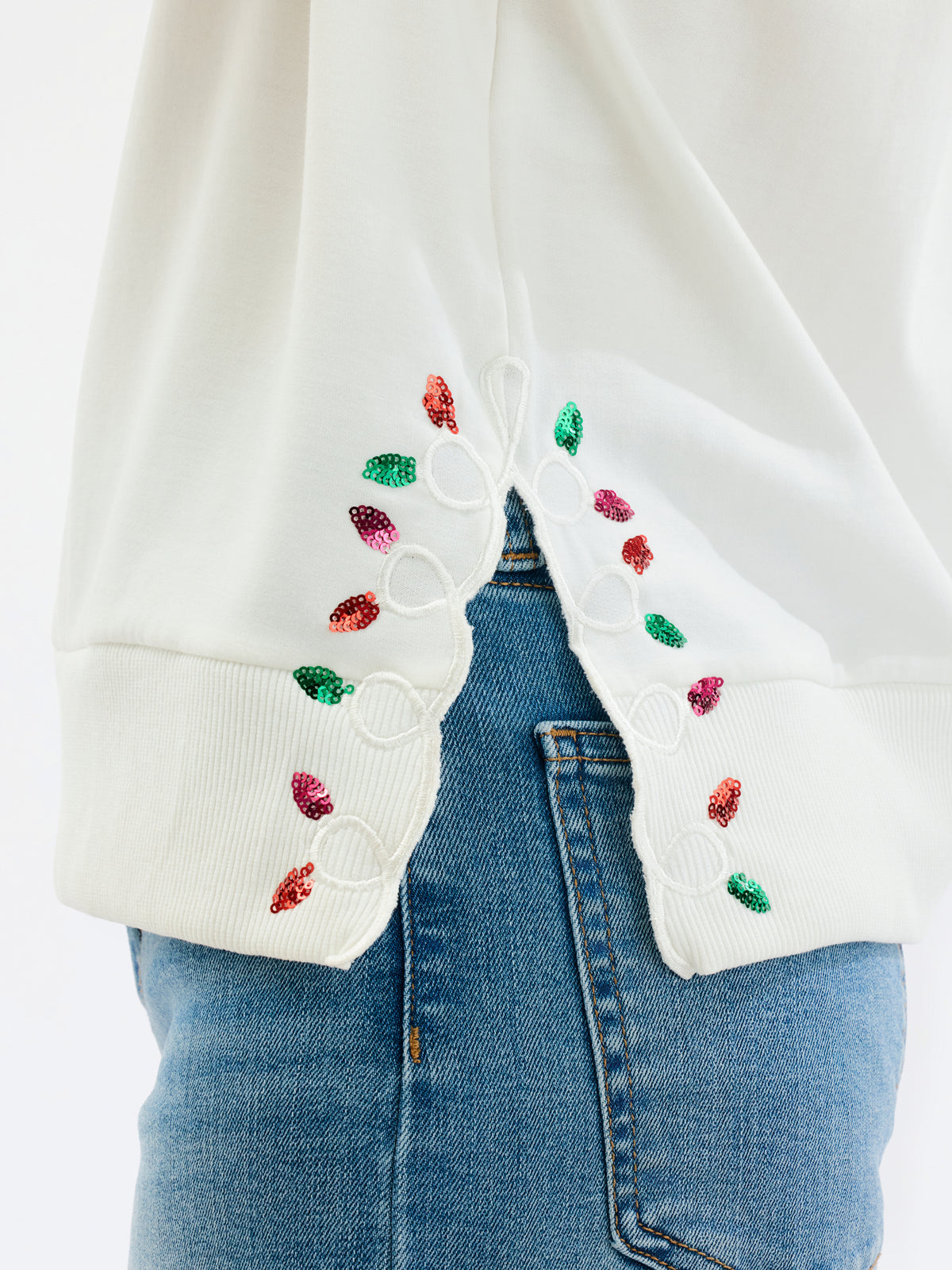 A woman wearing a cream-colored crewneck sweatshirt with 'Merry Bright Joyful' written in red and green glitter-style lettering on the front, paired with blue jeans, against a plain white studio background.