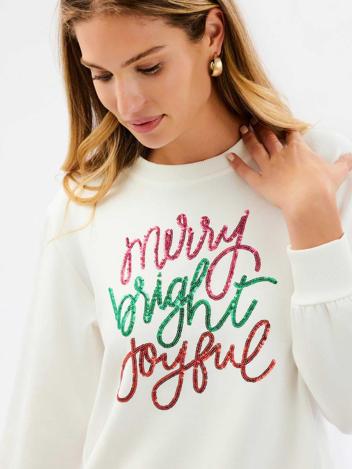 A woman wearing a cream-colored crewneck sweatshirt with 'Merry Bright Joyful' written in red and green glitter-style lettering on the front, paired with blue jeans, against a plain white studio background.