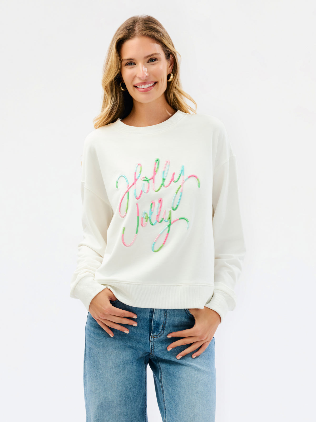 A woman wearing a cream-colored crewneck sweatshirt with 'Holly Jolly' written in a colorful pastel script across the chest, paired with blue jeans, against a plain white background.