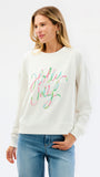 A woman wearing a cream-colored crewneck sweatshirt with 'Holly Jolly' written across the chest in a colorful, cursive script. She is paired with blue jeans, standing against a clean white studio background.