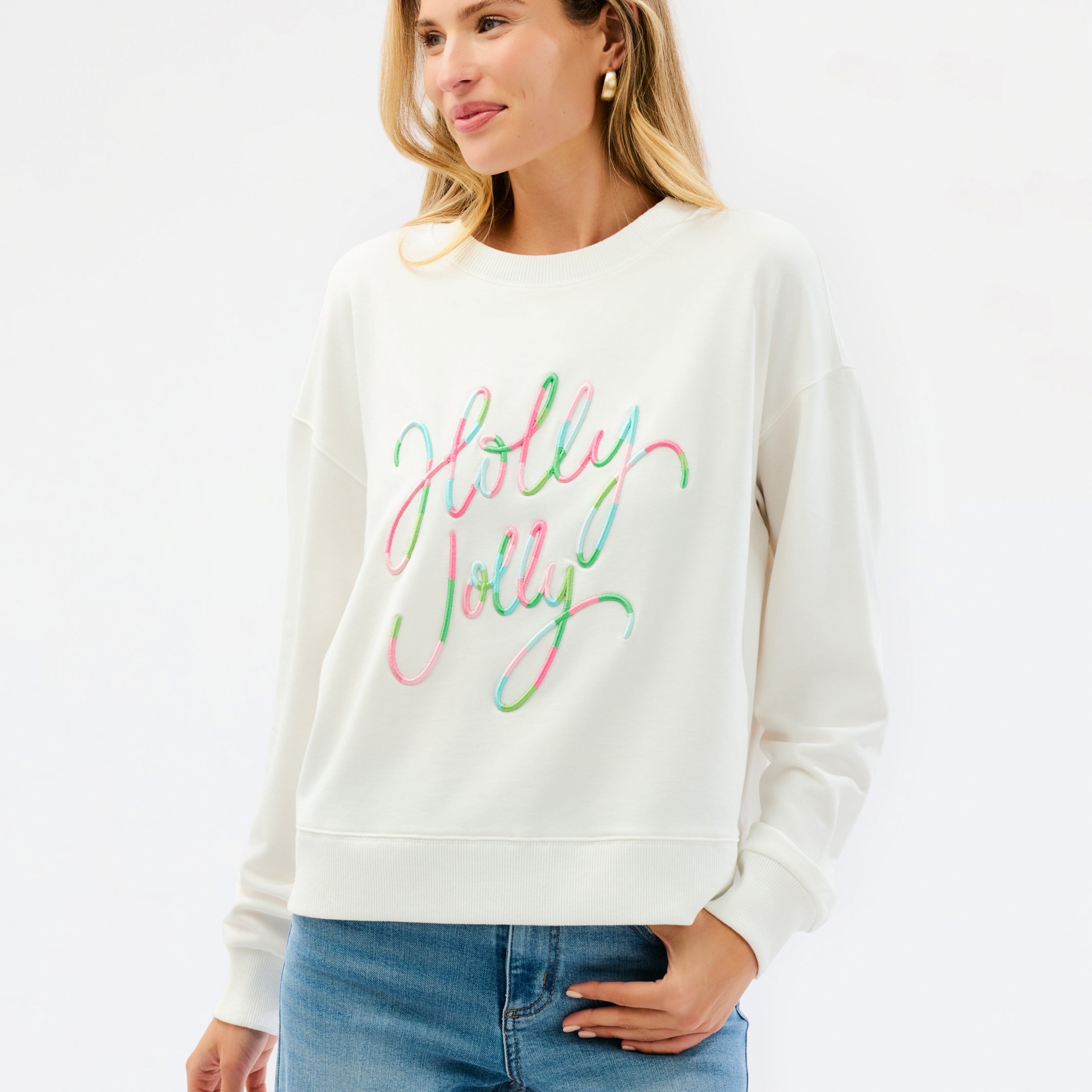 A woman wearing a cream-colored crewneck sweatshirt with 'Holly Jolly' written across the chest in a colorful, cursive script. She is paired with blue jeans, standing against a clean white studio background.
