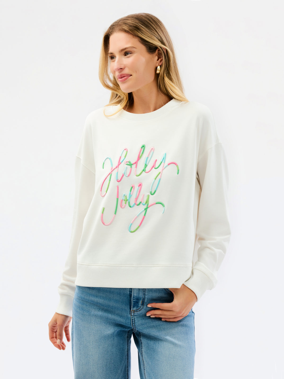 A woman wearing a cream-colored crewneck sweatshirt with 'Holly Jolly' written across the chest in a colorful, cursive script. She is paired with blue jeans, standing against a clean white studio background.