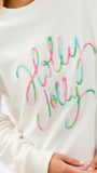 A close-up of a person wearing a cream-colored crewneck sweatshirt with 'Holly Jolly' embroidered in a cursive script across the chest, where the lettering is outlined in pink, green, and light blue.