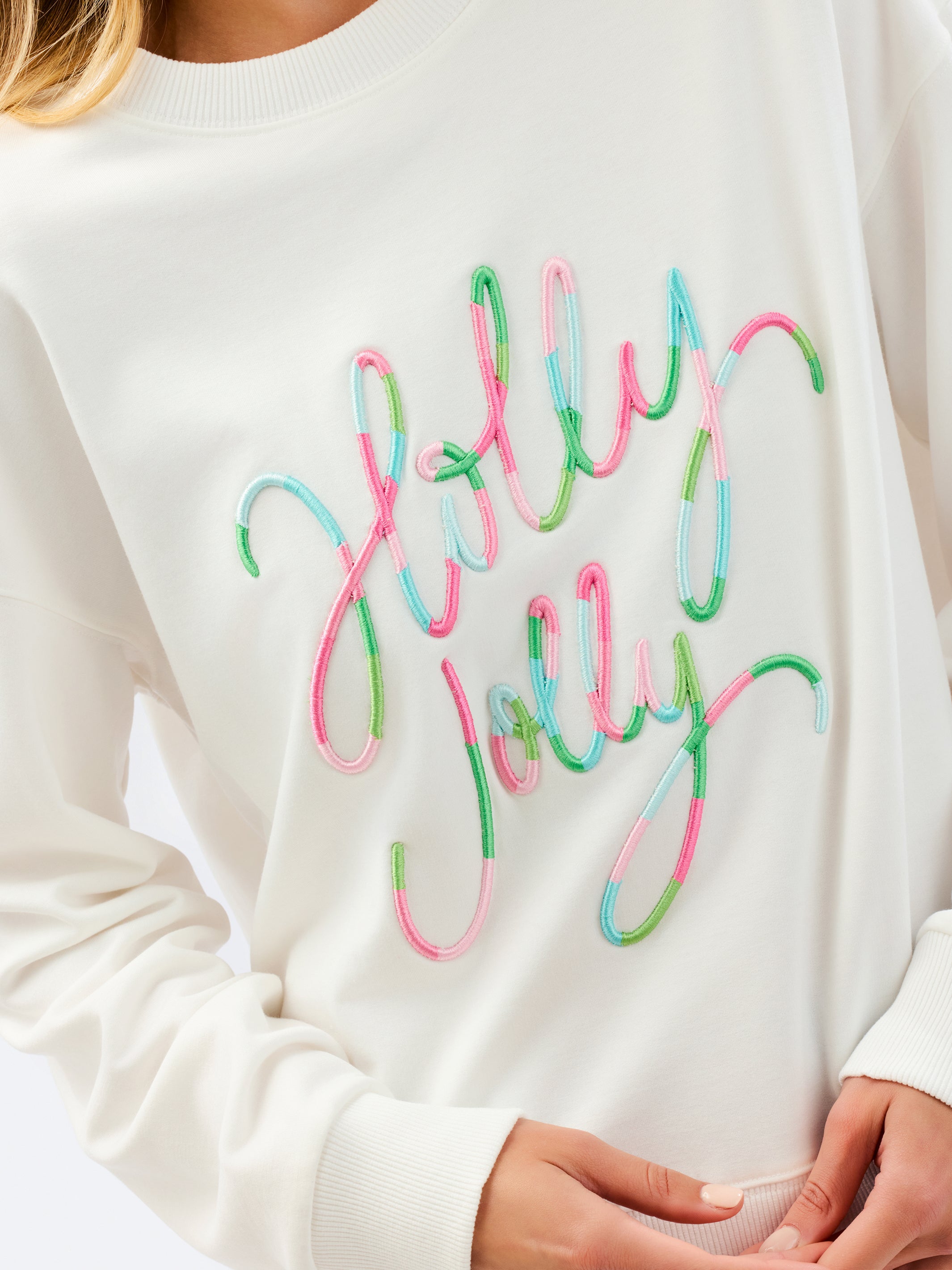 A close-up of a person wearing a cream-colored crewneck sweatshirt with 'Holly Jolly' embroidered in a cursive script across the chest, where the lettering is outlined in pink, green, and light blue.