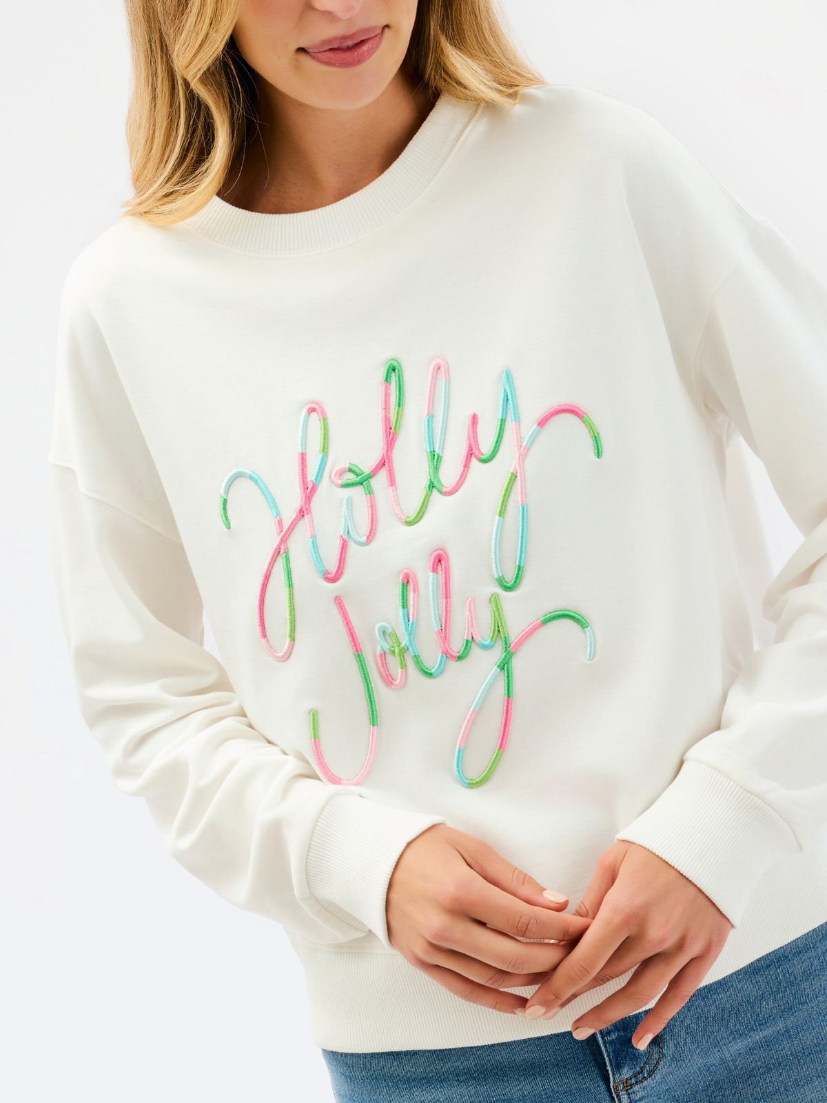 A woman wearing a cream-colored crewneck sweatshirt with 'Holly Jolly' written across the front in a colorful, neon-script font.