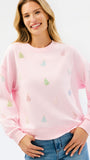 A woman wearing a light pink crewneck sweatshirt decorated with small multicolored Christmas tree motifs, paired with blue jeans against a plain white background.