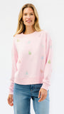 A woman wearing a light pink crewneck sweatshirt with small multicolored embroidered motifs, paired with blue jeans, against a plain white studio background.