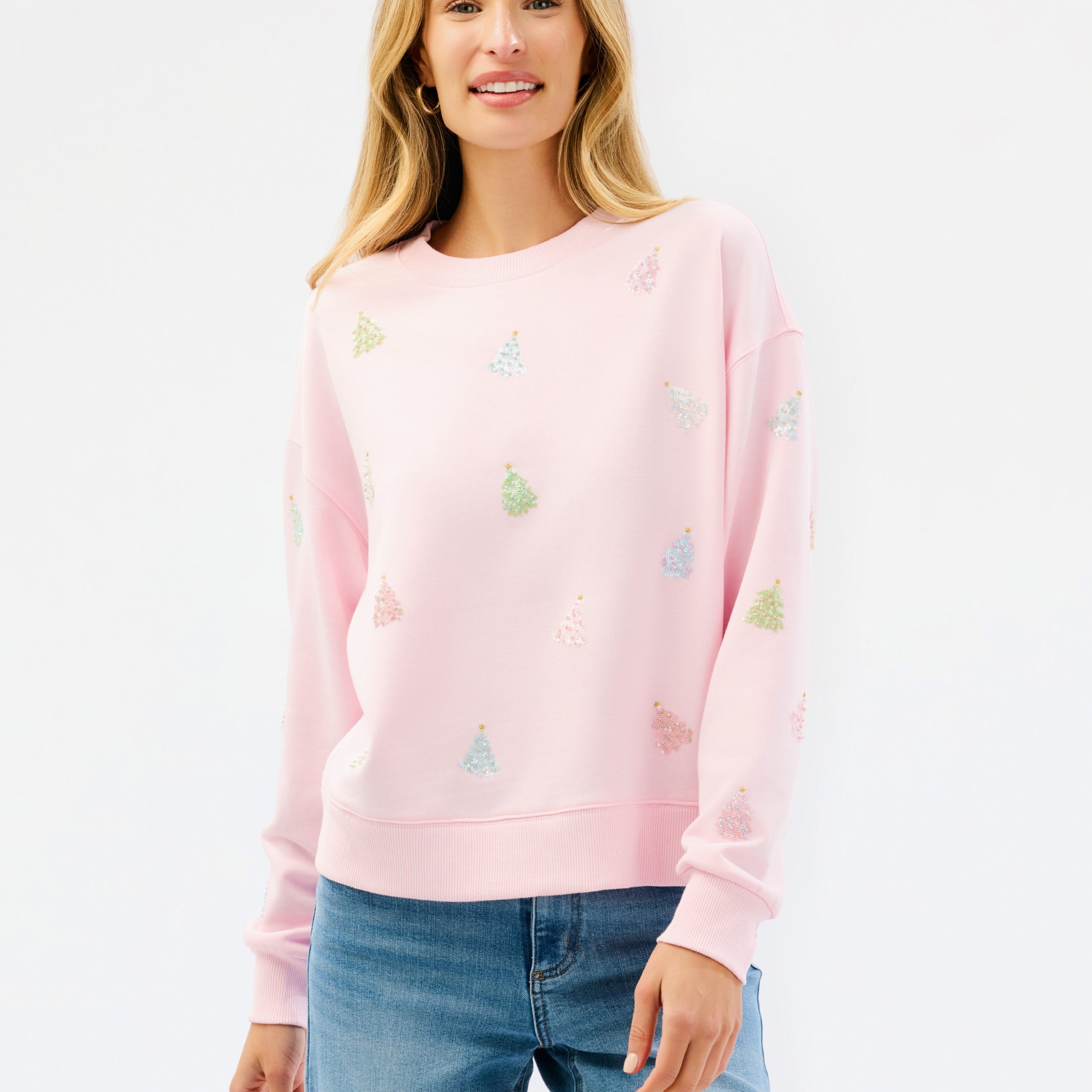 A woman wearing a light pink crewneck sweatshirt with small multicolored embroidered motifs, paired with blue jeans, against a plain white studio background.
