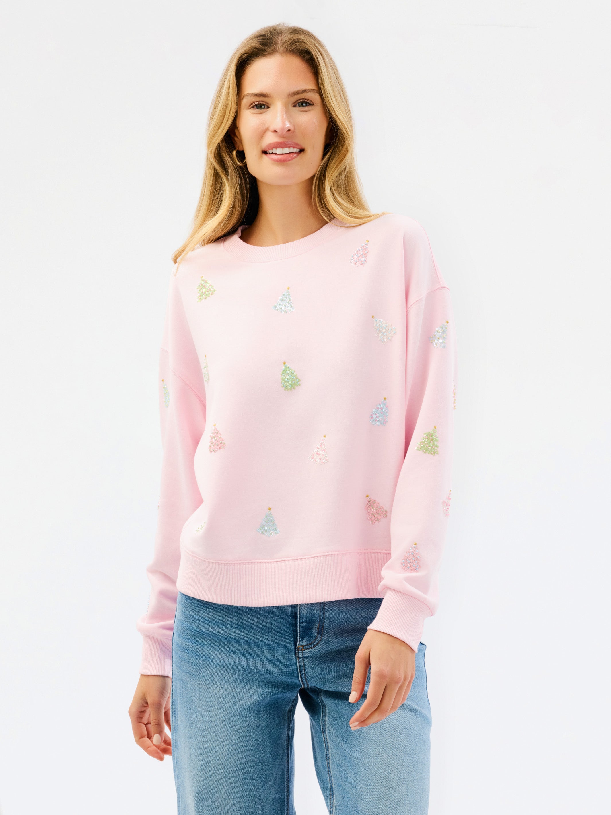 A woman wearing a light pink crewneck sweatshirt with small multicolored embroidered motifs, paired with blue jeans, against a plain white studio background.