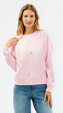 A woman modeling a light pink crewneck sweatshirt with small multicolored embroidered Christmas tree motifs, paired with blue jeans against a plain white studio background.