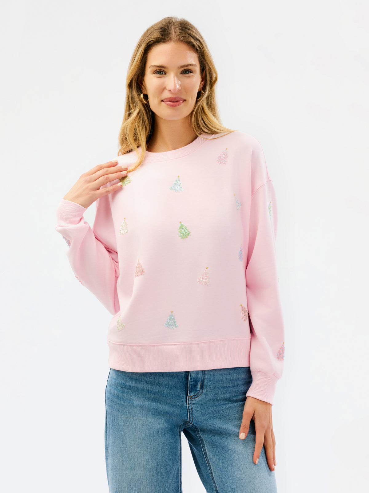 A woman modeling a light pink crewneck sweatshirt with small multicolored embroidered Christmas tree motifs, paired with blue jeans against a plain white studio background.