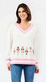 A woman modeling a white V-neck sweater with pink trim and colorful nutcracker graphics across the lower front, paired with blue jeans against a plain white background.