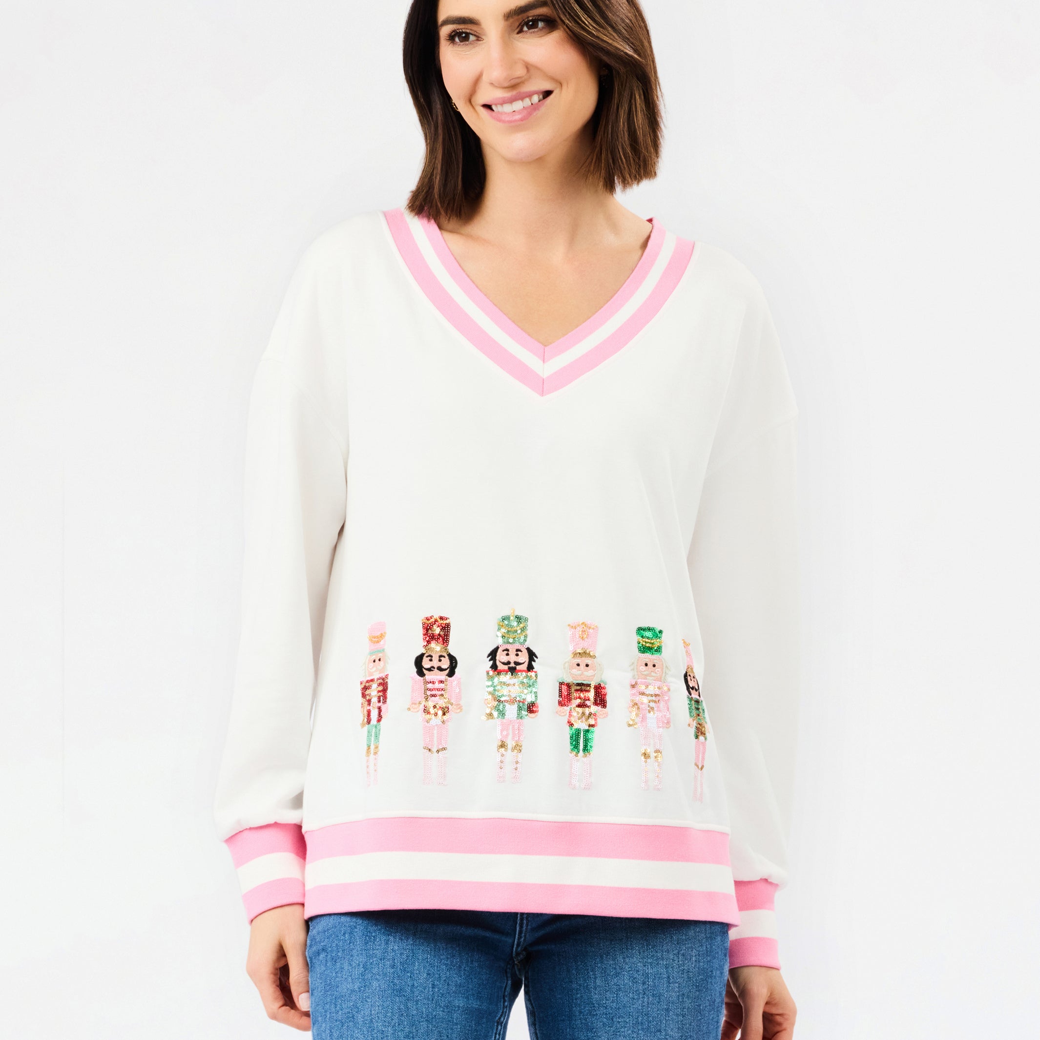 A woman modeling a white V-neck sweater with pink trim and colorful nutcracker graphics across the lower front, paired with blue jeans against a plain white background.