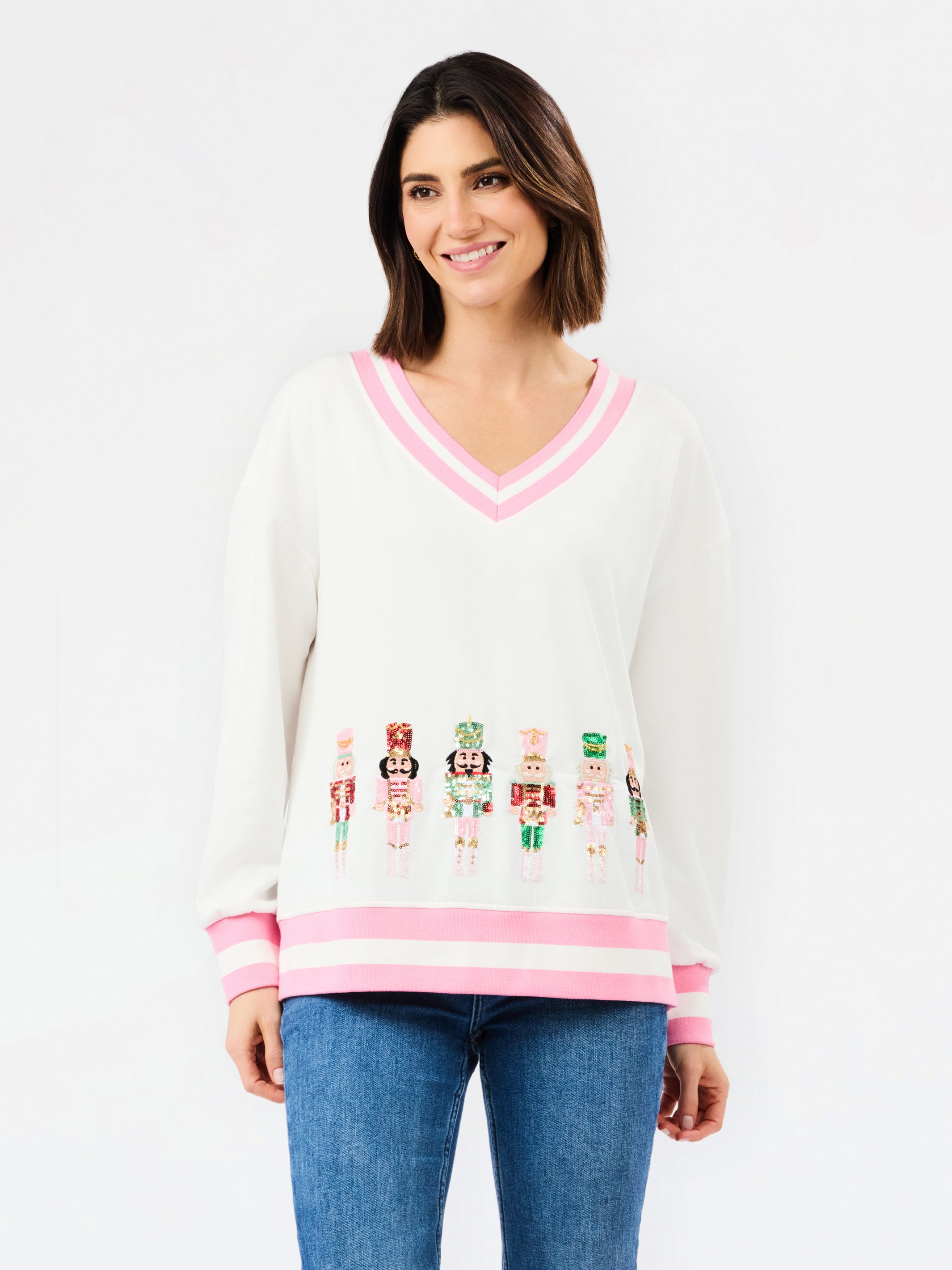 A woman modeling a white V-neck sweater with pink trim and colorful nutcracker graphics across the lower front, paired with blue jeans against a plain white background.