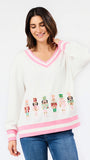 A woman wearing a white V-neck sweater with pink trim and embroidered nutcracker figures, paired with blue jeans, against a plain white background.