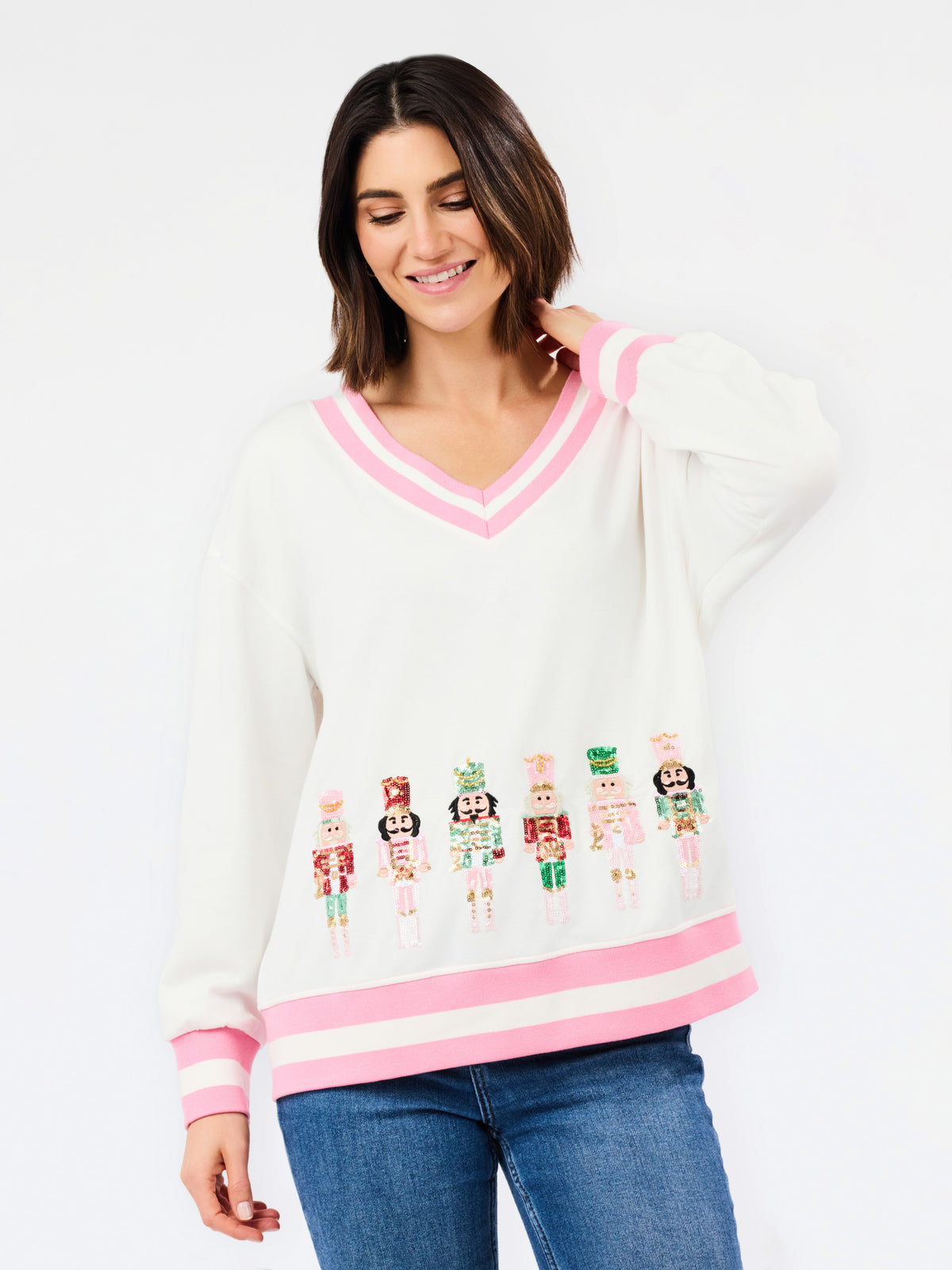 A woman wearing a white V-neck sweater with pink trim and embroidered nutcracker figures, paired with blue jeans, against a plain white background.