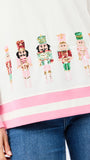A close-up of a white sweatshirt with pink ribbed trim and a row of colorful sequined nutcracker figures across the chest, worn with blue jeans.