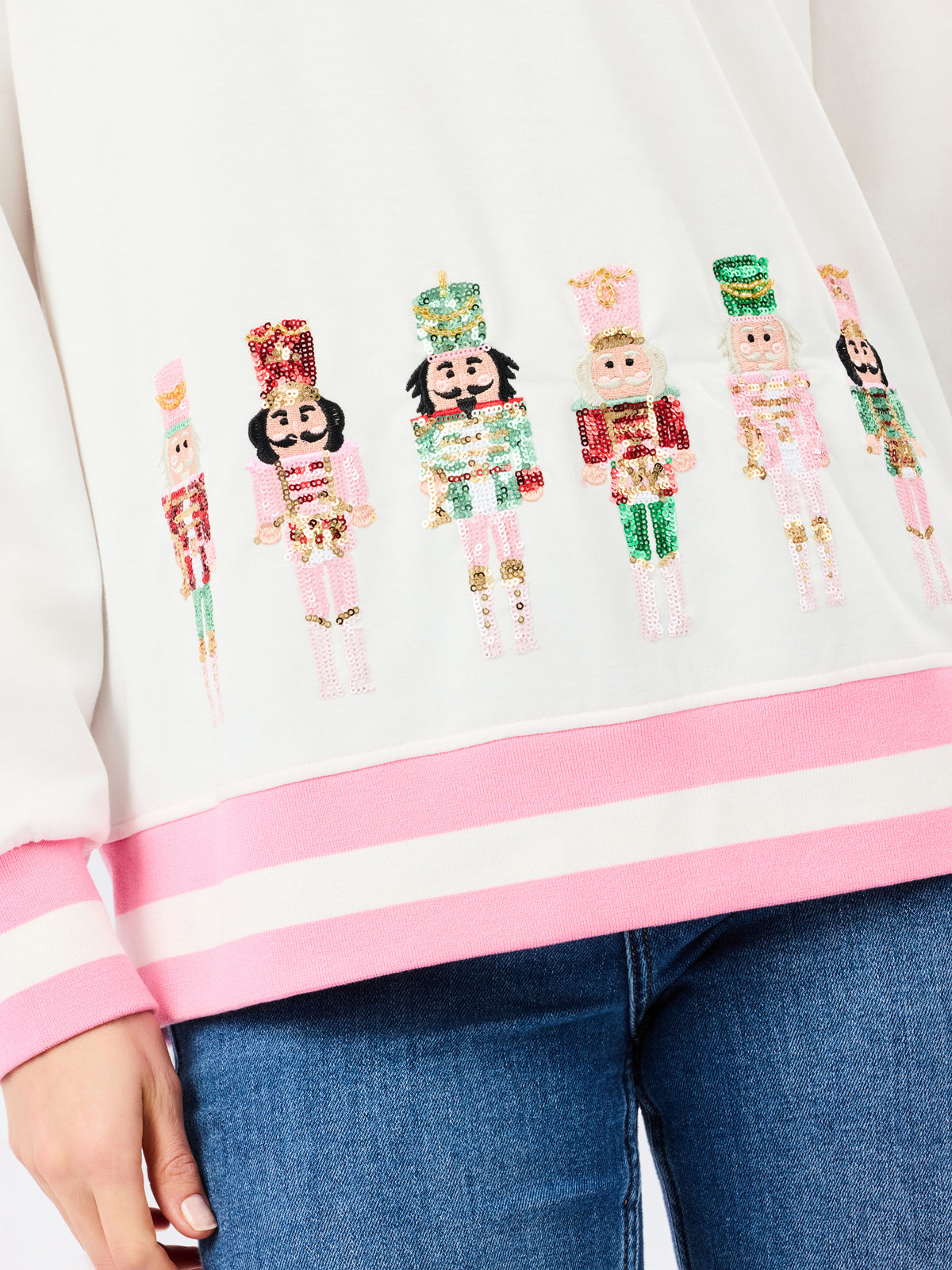 A close-up of a white sweatshirt with pink ribbed trim and a row of colorful sequined nutcracker figures across the chest, worn with blue jeans.