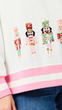 Close-up of a white sweatshirt with pink striped ribbed cuffs and hem, featuring a row of sequined nutcracker figures across the chest, worn with blue jeans.