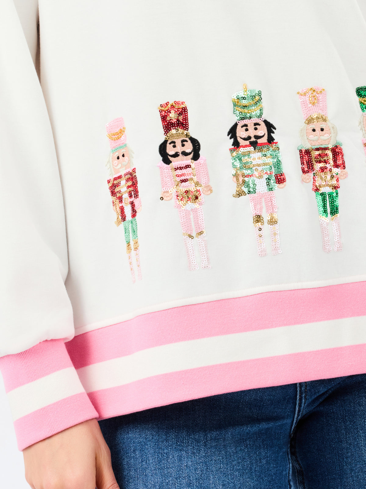 Close-up of a white sweatshirt with pink striped ribbed cuffs and hem, featuring a row of sequined nutcracker figures across the chest, worn with blue jeans.