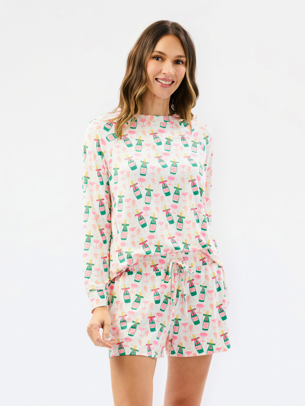 A woman modeling a matching long-sleeve top and shorts set in a white fabric with a pink and green pattern of wine bottles, set against a plain white background.