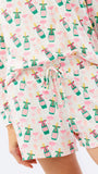 A close-up of a person wearing a matching long-sleeve top and shorts set with a white background and an all-over print of pink champagne bottles and green flutes, tied with a white drawstring.