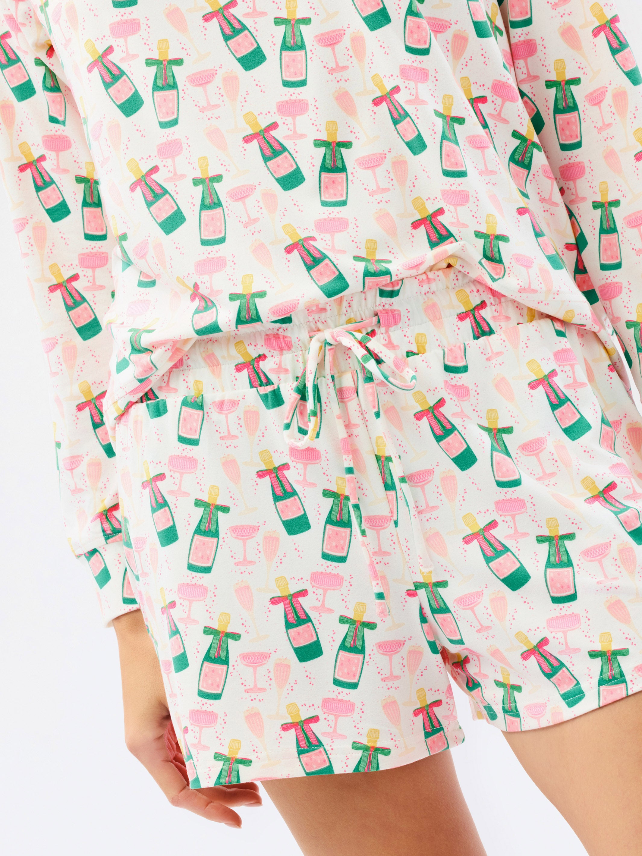 A close-up of a person wearing a matching long-sleeve top and shorts set with a white background and an all-over print of pink champagne bottles and green flutes, tied with a white drawstring.