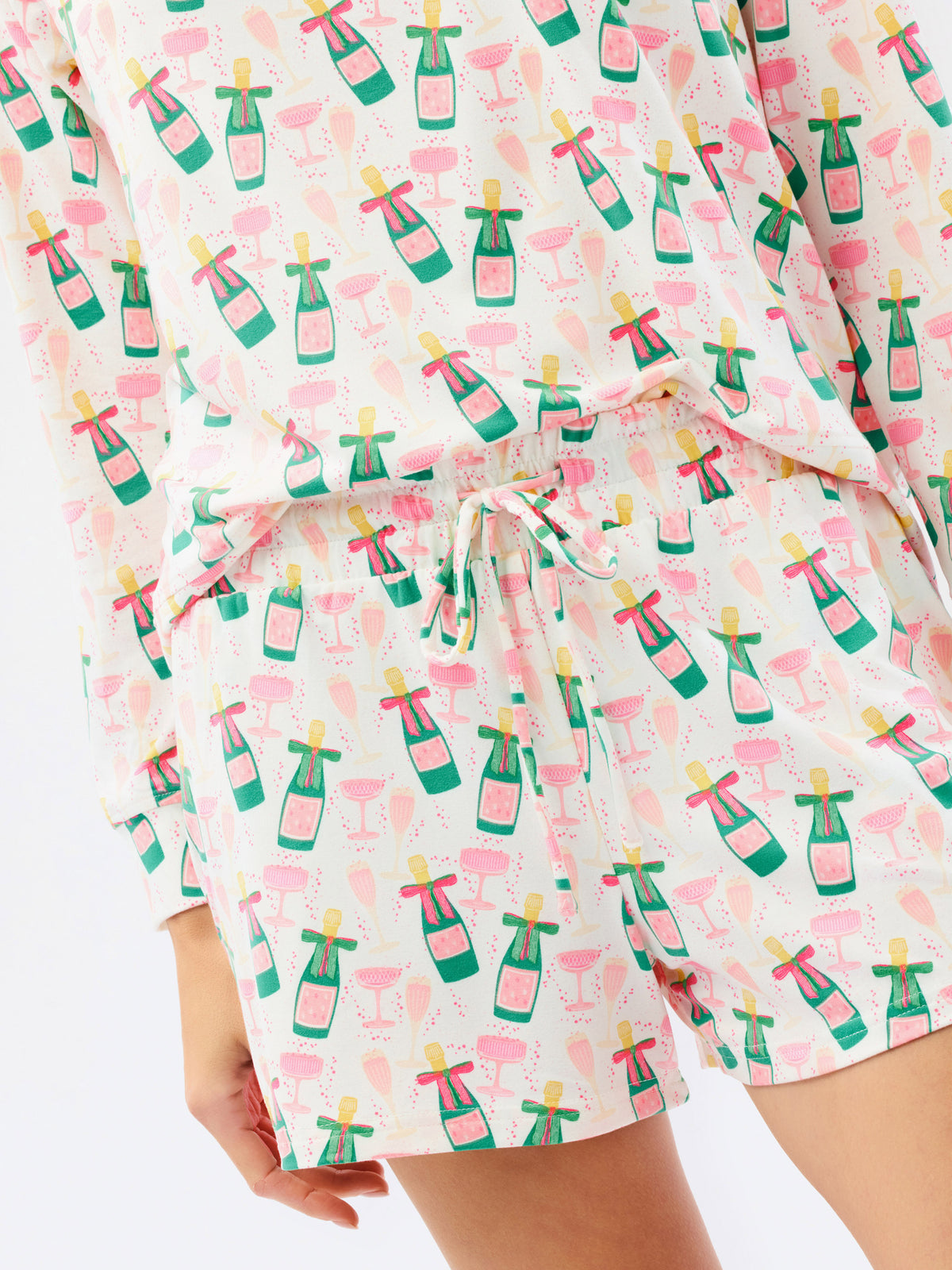 A close-up of a person wearing a matching long-sleeve top and shorts set with a white background and an all-over print of pink champagne bottles and green flutes, tied with a white drawstring.