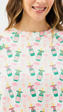A close-up of a woman wearing a white short-sleeve top with a repeating pattern of green champagne bottles, pink cocktail glasses, and yellow straws.
