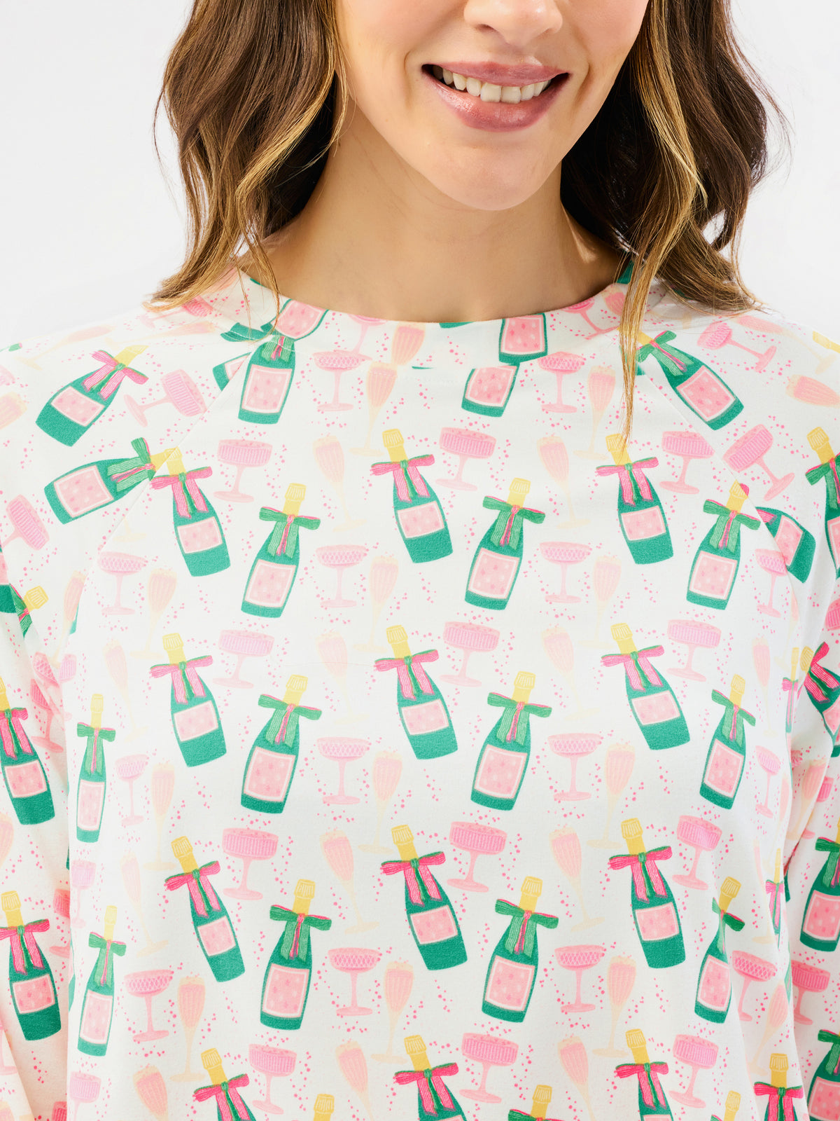 A close-up of a woman wearing a white short-sleeve top with a repeating pattern of green champagne bottles, pink cocktail glasses, and yellow straws.