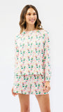 A woman modeling a matching long-sleeve top and shorts pajama set with a pink and white tropical print featuring palm trees and drinks, against a plain white studio background.