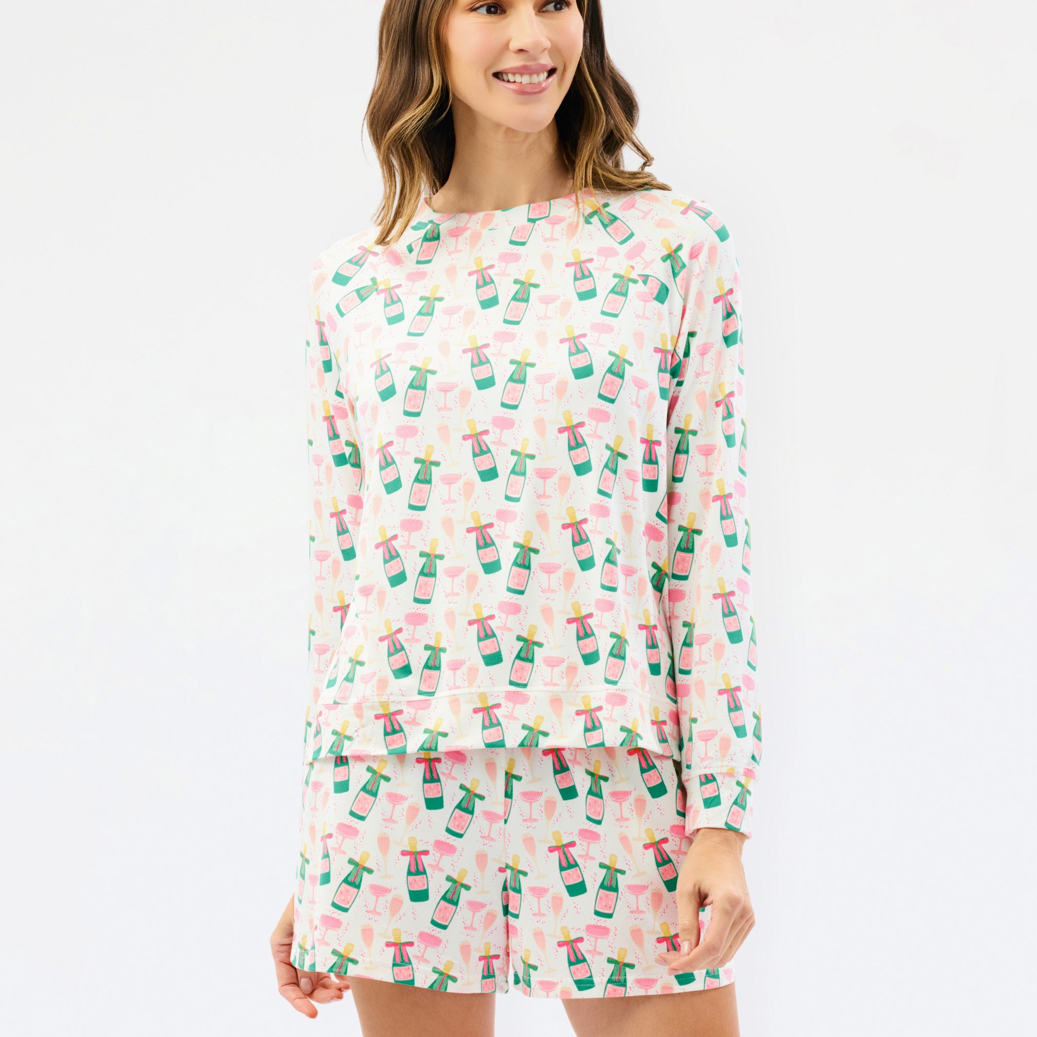 A woman modeling a matching long-sleeve top and shorts pajama set with a pink and white tropical print featuring palm trees and drinks, against a plain white studio background.