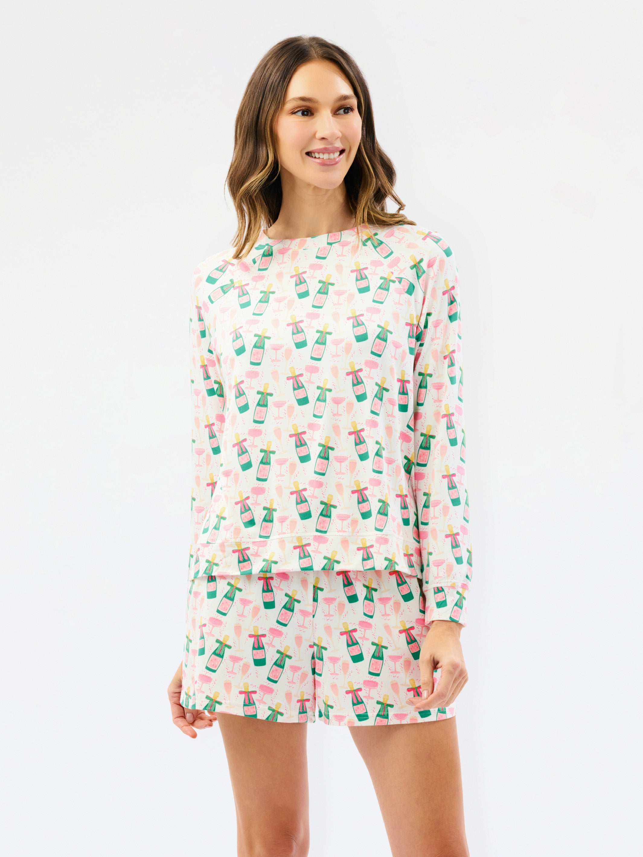 A woman modeling a matching long-sleeve top and shorts pajama set with a pink and white tropical print featuring palm trees and drinks, against a plain white studio background.
