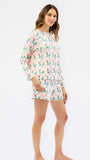 A woman modeling a long-sleeve, tiered sleepwear set with a pink flamingo and green palm tree pattern on a white background, shot against a seamless white studio backdrop.