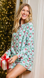 A woman wearing a light blue-green long-sleeve pajama romper with a green wreath and red bow holiday print, leaning against a cream door next to a lit Christmas tree and holding a wrapped gift box.