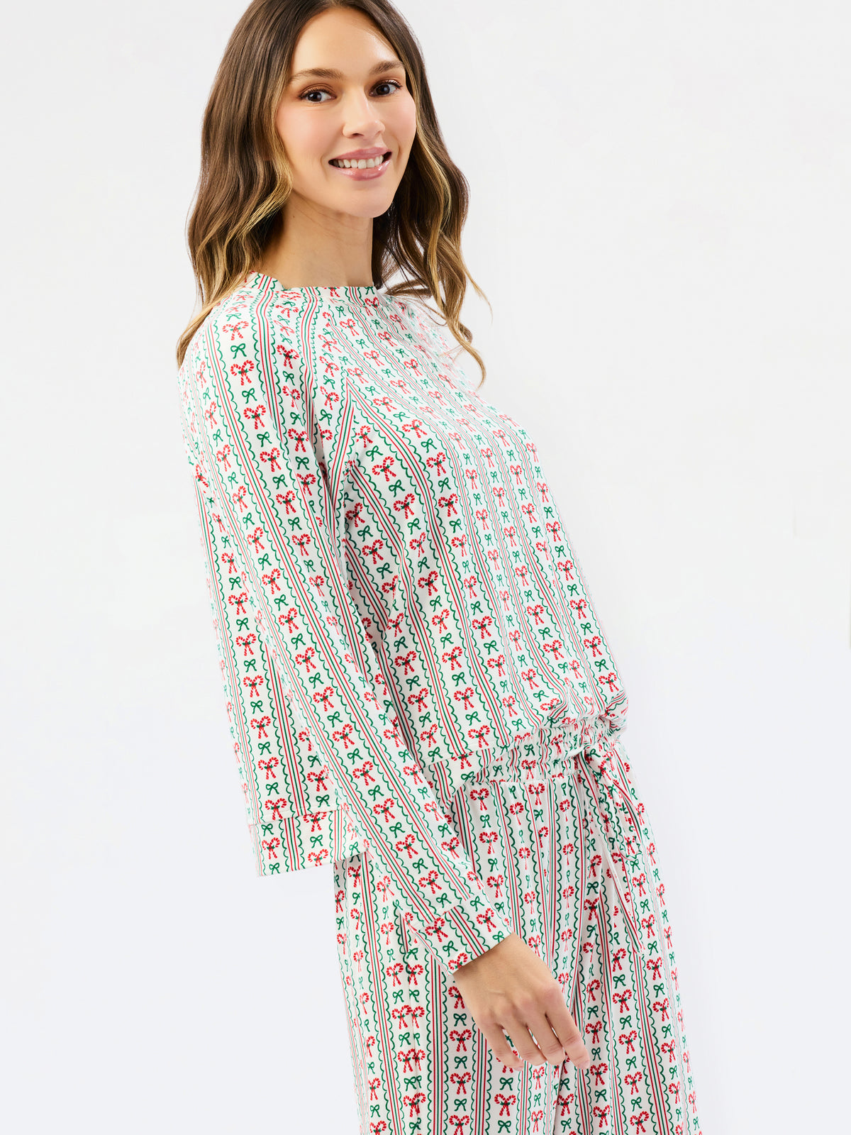A woman wearing a long-sleeve jumpsuit with a festive red and green holiday pattern on a white background, photographed against a clean white studio backdrop.