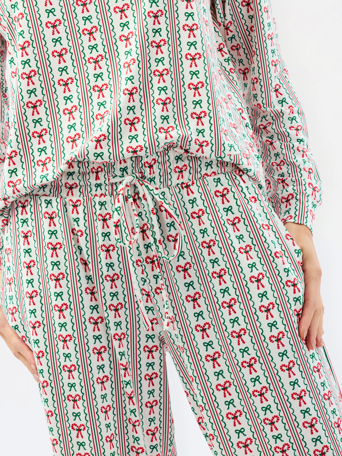 A person wearing a matching long-sleeve top and pants set with a festive Christmas print of red bows, green ribbons, and vertical stripes on a white background.