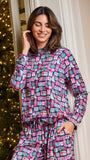 A woman wearing a matching long-sleeve pajama set with a colorful Christmas gift box print, standing in front of a decorated Christmas tree and cream-colored paneled wall.