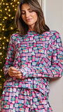 A woman wearing a long-sleeve high-neck pajama top and matching pants with a colorful Christmas gift box pattern, posed in front of a decorated Christmas tree.