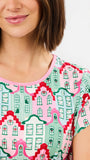 A close-up of a woman wearing a short-sleeve top with a colorful architectural house pattern in pink, green, and red on a light background.